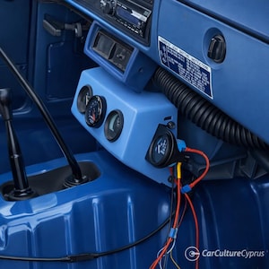 Può includere: Un cruscotto blu di un veicolo con un supporto personalizzato per tre indicatori analogici circolari, un indicatore separato montato a destra, cavi rossi e neri a vista, una leva del cambio nera e un adesivo con le istruzioni per il cambio.