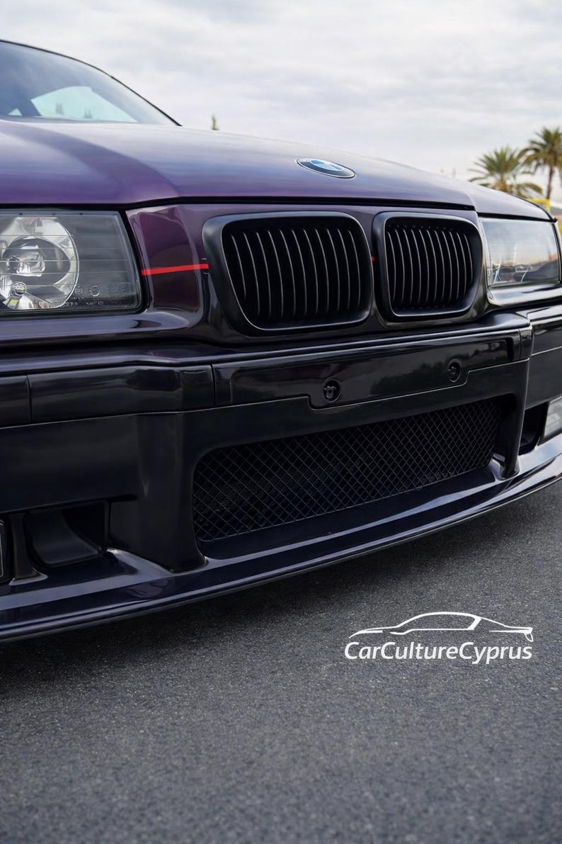 May include: A dark purple BMW car front end featuring a black kidney grille, a lower mesh air intake, and clear headlights. A thin red horizontal stripe accents the hood near the grille. The text CarCultureCyprus is visible in white.
