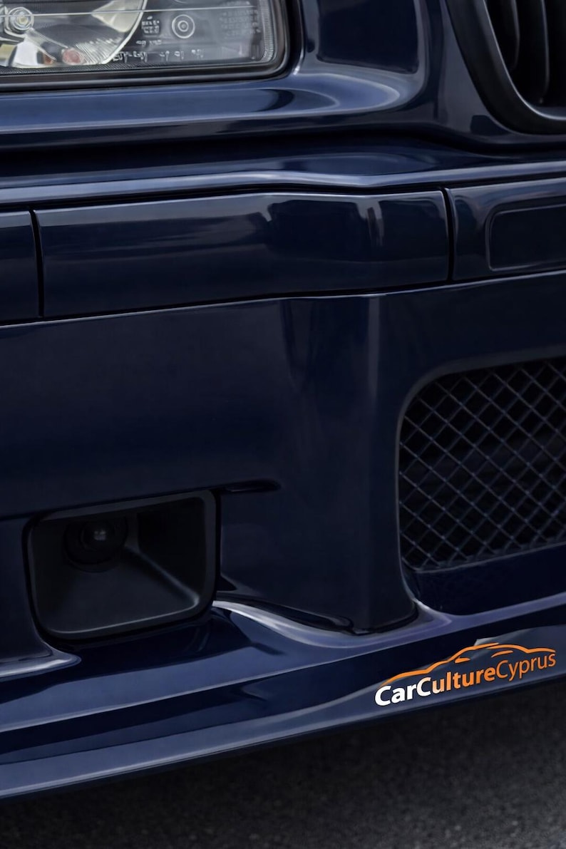May include: A dark blue car bumper featuring a black mesh grille, a rectangular fog light housing, and a headlight assembly at the top. The text CarCultureCyprus is printed in white and orange on the lower right side of the bumper surface.