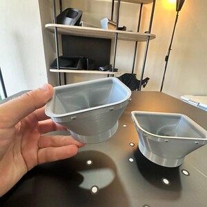 May include: Two light gray, funnel-shaped plastic components with a wide, square-like opening that tapers down to a circular base. Each piece features a flat rim with small, evenly spaced rectangular tabs along the outer edge of the top opening.