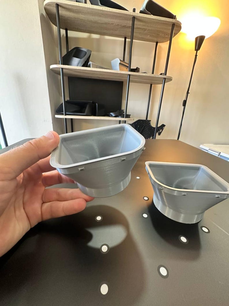 May include: Two light gray, funnel-shaped plastic components with a wide, square-like opening that tapers down to a circular base. Each piece features a flat rim with small, evenly spaced rectangular tabs along the outer edge of the top opening.