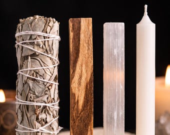 Può includere: Un mazzetto di foglie di salvia essiccate legate con uno spago bianco, un pezzo rettangolare di legno marrone, una bacchetta di cristallo di selenite bianca traslucida e una sottile candela bianca disposti in fila su uno sfondo scuro.