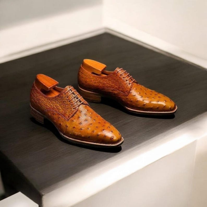 Op de afbeelding: Een paar bruine leren herenschoenen met een gestructureerd struisvogelpatroon. De schoenen hebben een klassiek vetersluiting ontwerp, een lage hak en worden getoond met houten schoenspanners. De schoenen staan op een donkere ondergrond.