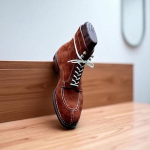 Op de afbeelding: Een bruine suède laars met witte veters en stiksels. De laars staat op een houten oppervlak, wat het ontwerp benadrukt. De laars heeft een klassieke stijl, geschikt voor diverse gelegenheden. De laars is ongeveer 25 cm lang.