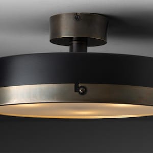 May include: A contemporary ceiling light. It features a black cylindrical shade with a brushed brass band. The fixture has a matching brass base and emits a warm, diffused light. The design is minimalist and elegant.