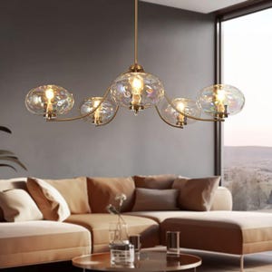 Peut inclure: Un lustre moderne avec six globes en verre irisé, chacun contenant une lumière chaude. Le luminaire a une monture en métal doré et est suspendu au plafond. Le lustre est au-dessus d'un canapé beige.