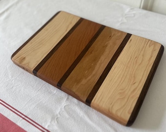 May include: A rectangular wooden cutting board with a striped design. The board is made of light and dark wood, creating a visually appealing contrast. The board is perfect for serving cheese, charcuterie, or other appetizers.