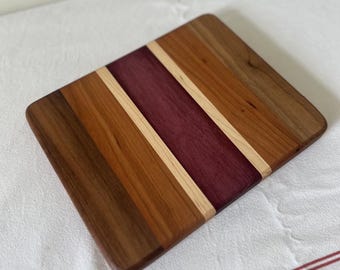 May include: A rectangular wooden cutting board with a striped design. The board features alternating stripes of various wood tones, including dark brown, reddish-brown, light tan, and deep purple. The edges are rounded.