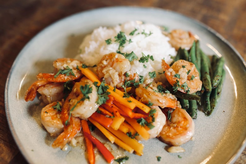 May include: A plate of cooked shrimp with rice, green beans, and colorful julienned vegetables. The shrimp are golden brown and garnished with fresh parsley. The dish is served on a light green plate.