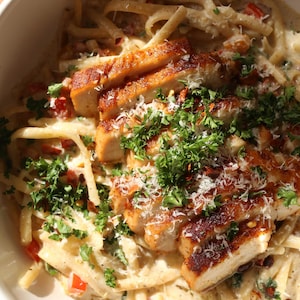 Op de afbeelding: Een kom romige pasta met gesneden, aangebraden kipfilet. De pasta is bedekt met een witte saus met rode paprika's en kruiden. Gegarneerd met verse peterselie en geraspte kaas.