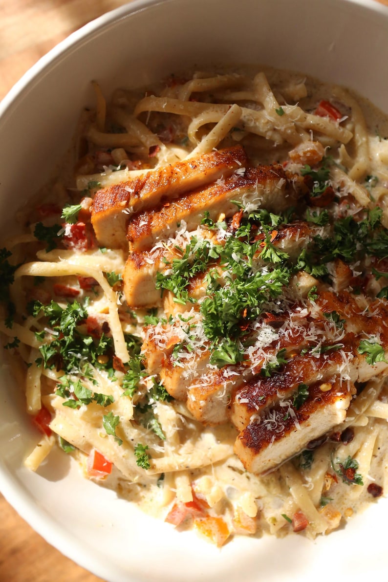 Op de afbeelding: Een kom romige pasta met gesneden, aangebraden kipfilet. De pasta is bedekt met een witte saus met rode paprika's en kruiden. Gegarneerd met verse peterselie en geraspte kaas.