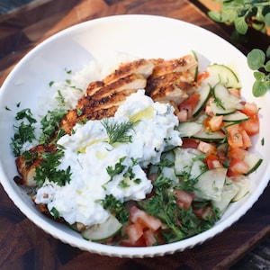 May include: A white bowl filled with a colorful meal. The bowl contains grilled chicken, white rice, a cucumber and tomato salad, and a dollop of creamy white sauce, garnished with fresh herbs. The food is on a wooden surface.