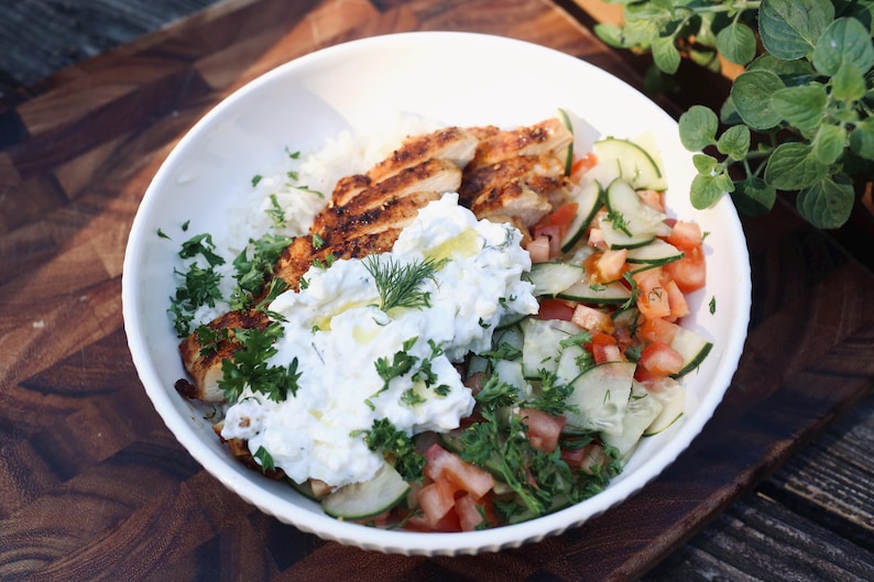 May include: A white bowl filled with a colorful meal. The bowl contains grilled chicken, white rice, a cucumber and tomato salad, and a dollop of creamy white sauce, garnished with fresh herbs. The food is on a wooden surface.