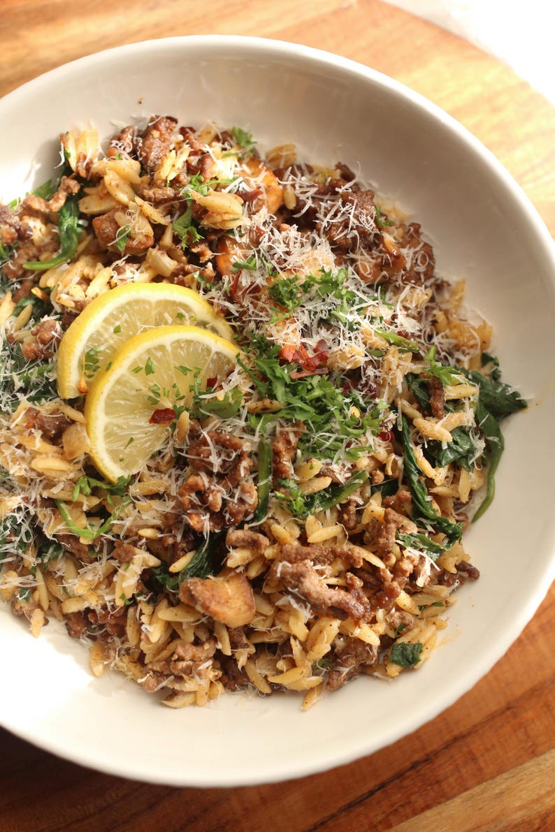 Op de afbeelding: Een witte kom gevuld met een hartig gerecht met orzo pasta, gehakt, spinazie en citroenschijfjes. Het gerecht is gegarneerd met verse peterselie en geraspte kaas, wat een visueel aantrekkelijke maaltijd cre&euml;ert.