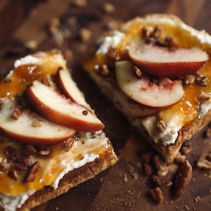 May include: Two slices of a pastry topped with white cream, peach slices, and chopped nuts. A drizzle of orange sauce adds a glossy finish. The food is presented on a dark wooden surface, with scattered nuts.