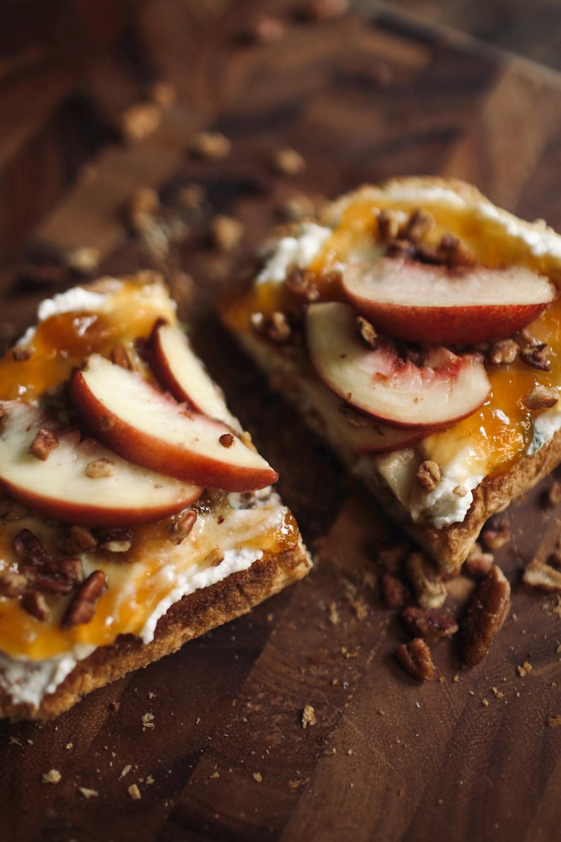 May include: Two slices of a pastry topped with white cream, peach slices, and chopped nuts. A drizzle of orange sauce adds a glossy finish. The food is presented on a dark wooden surface, with scattered nuts.