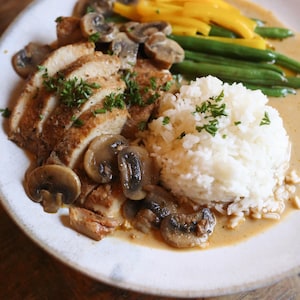 May include: A plate of food featuring sliced chicken with a mushroom sauce, sauteed mushrooms, white rice, green beans, and yellow squash. Garnished with fresh parsley. The meal is served on a white plate.