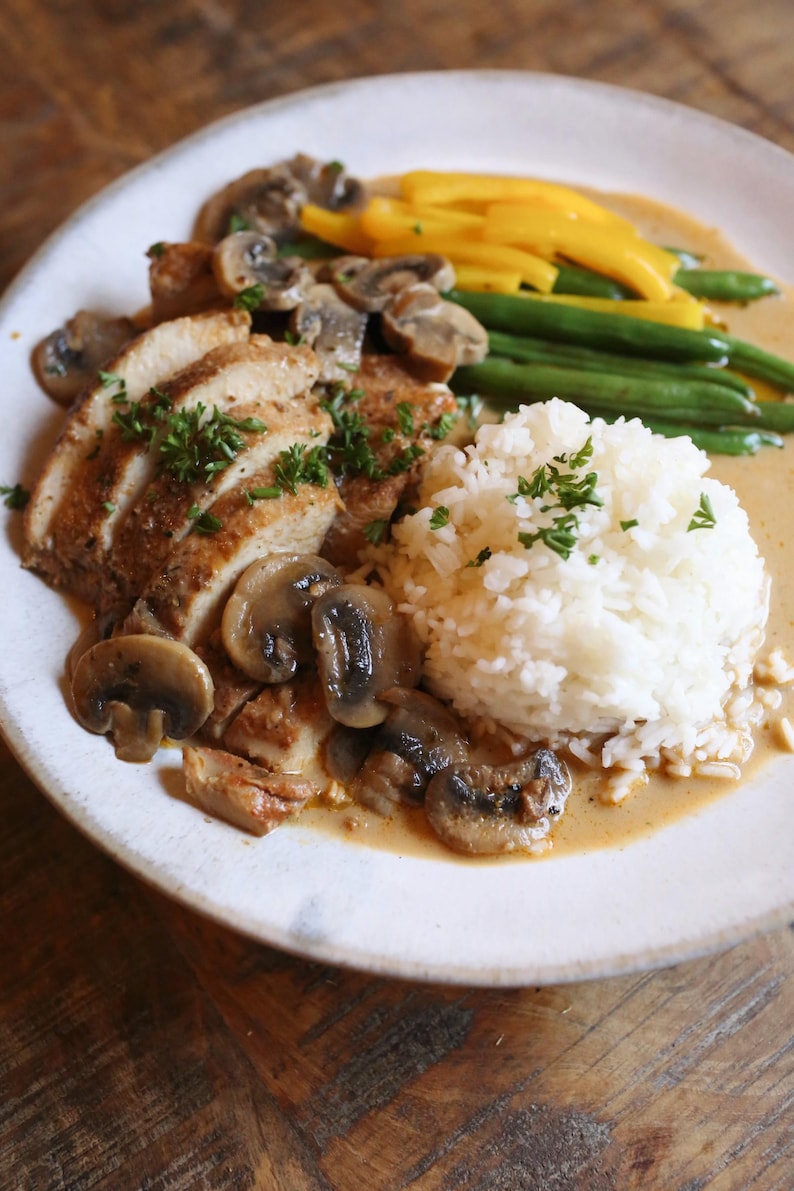 May include: A plate of food featuring sliced chicken with a mushroom sauce, sauteed mushrooms, white rice, green beans, and yellow squash. Garnished with fresh parsley. The meal is served on a white plate.