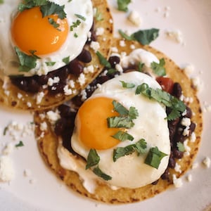 May include: Two fried eggs with bright orange yolks sit atop tortillas with black beans, white cheese, and cilantro. The dish is served on a white plate, with a rustic wooden table in the background.