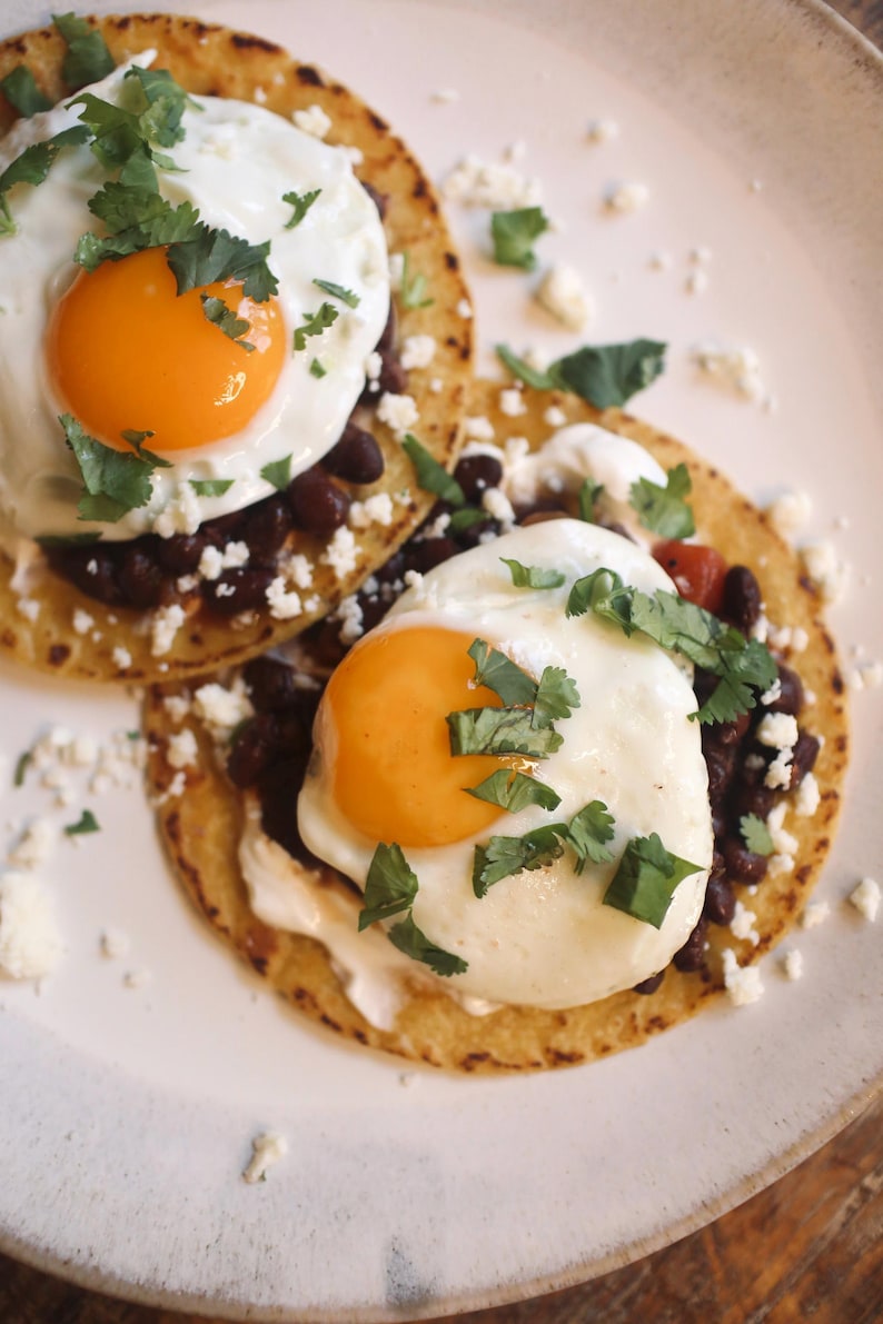 May include: Two fried eggs with bright orange yolks sit atop tortillas with black beans, white cheese, and cilantro. The dish is served on a white plate, with a rustic wooden table in the background.