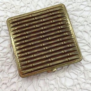 May include: A vintage gold-toned compact with a bamboo-style textured design. The rectangular compact has a hinged closure and a detailed, repeating pattern across its surface. The compact is likely made of metal and shows signs of age.