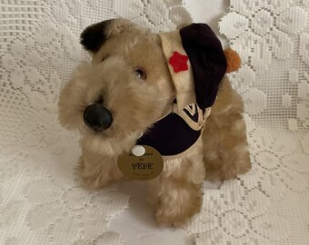 May include: A tan plush toy dog wearing a blue and white sailor hat with a red star. The dog has a black nose and brown eyes. The dog is wearing a blue and white sailor collar. The dog has a tag that says "Pepe".