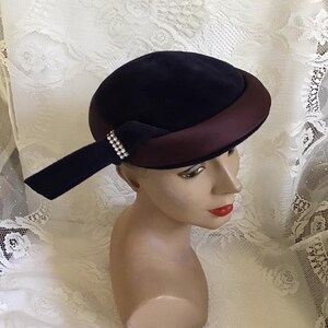 May include: A dark blue velvet hat with a burgundy satin trim and a rhinestone embellishment. The hat is perched on a mannequin head.