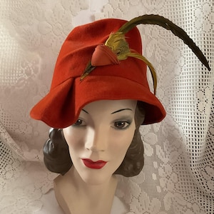 May include: A vintage orange cloche hat with a decorative feather and ribbon accent. The hat is displayed on a mannequin head. The hat's brim is slightly flared, and the feather is a mix of yellow and brown.