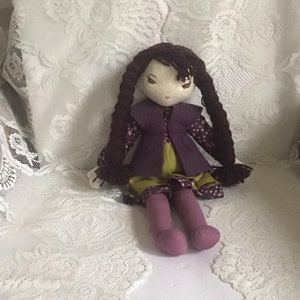 May include: A handmade doll with brown hair in braids, wearing a purple vest, a green skirt, and pink tights. The doll is sitting on a white lace surface.