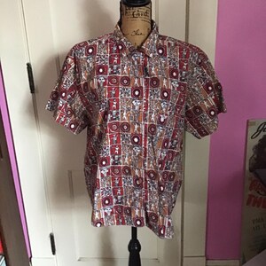 May include: A short-sleeved button-down shirt with a colorful geometric pattern. The pattern features stylized figures and circles in shades of red, brown, white, and tan.