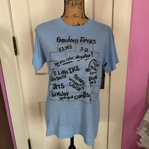 May include: Light blue t-shirt with a black graphic print of a brick wall with the text "Fabulous Fifties", "Elvis 3-0", "See you later, alligator", "I Like Ike", "Aloha Skirts", "Rebel Without a Cause", "Duck Tails", "Jets", "Bill Haley and the Comets".