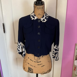 May include: A navy blue cropped jacket with a white and black patterned collar and cuffs. The jacket has a button-up front and two flap pockets.