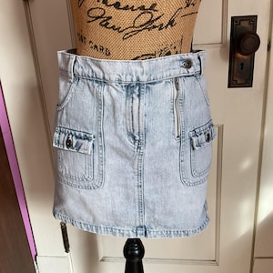 May include: A light blue denim skirt with a zip fastening and two large pockets on the front. The skirt is mini length and has a slightly distressed look.