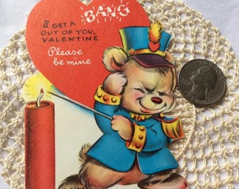 May include: A vintage Valentine's Day card featuring a cartoon bear dressed as a drummer, holding a drumstick and standing in front of a red dynamite stick with a lit fuse. The card is shaped like a heart and has the text "I get a BANG out of you, VALENTINE Please be mine" on it.