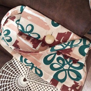 May include: A small, cream-colored fabric bag with a green and brown abstract pattern. The bag has a brown tassel and a wooden button closure.