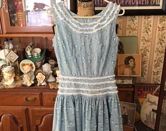 May include: A sleeveless, light blue vintage dress with a floral pattern and white lace trim. The dress is hanging on a hanger and has a fitted bodice and a flared skirt. The dress is approximately 40 inches long.