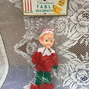 Può includere: Una decorazione da tavolo natalizia vintage con un elfo rosso e verde con un cappello bianco e un bordo di glitter dorato. L'elfo è confezionato in un sacchetto di plastica trasparente con un'etichetta del prezzo che recita "Christmas Table Decoration, Key 1 98-94 36c".