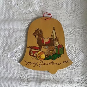 May include: A wooden bell ornament with a painted image of a rocking horse, a teddy bear, a toy drum, a ball, and a wrapped gift. The bell is painted brown and has the text "Merry Christmas 1981" written on it.