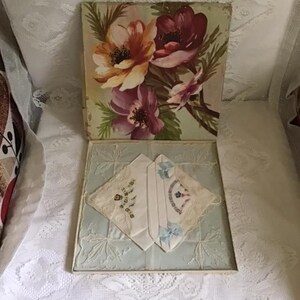 May include: A vintage handkerchief set in a floral box. The box lid features a painting of orange, purple, and pink flowers. Inside, two white handkerchiefs with embroidered details and blue bows rest on a light blue patterned base.
