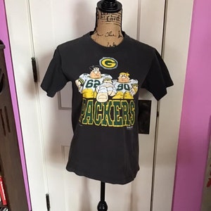 Könnte beinhalten: Schwarzes T-Shirt mit einem grünen und gelben Cartoon-Bild von drei Green Bay Packers Footballspielern. Das Shirt hat den Text "PACKERS" auf der Vorderseite.
