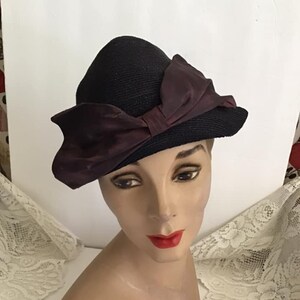 May include: A black straw hat with a large burgundy satin bow. The hat is perched on a mannequin head.