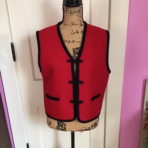 Pode incluir: Um colete de lã vermelho com acabamento preto e fechos de botão. O colete tem um decote em V e dois bolsos.