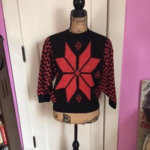 May include: A black and red knitted sweater with a large red snowflake design on the front. The sweater has a crew neck and long sleeves.