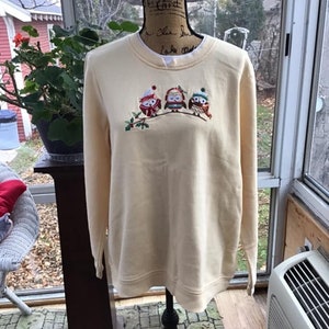 May include: A cream colored sweatshirt with a festive design of three owls wearing winter hats and scarves. The owls are embroidered on the front of the sweatshirt.