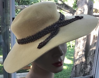 May include: A wide-brimmed hat made of felt in a pale yellow color. The hat is adorned with a brown beaded trim that wraps around the crown and ties in a bow at the front.