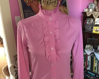 May include: A pink long-sleeved blouse with a ruffled collar and cuffs. The blouse has a button-up front and a fitted silhouette.