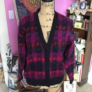 May include: A black cardigan sweater with a v-neck and button closure. The sweater has a plaid pattern in shades of pink, purple, blue, and black.