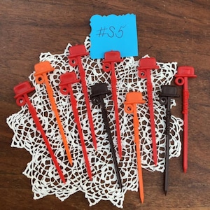 Peut inclure: Un ensemble de 12 porte-cure-dents en plastique vintage. Les porte-cure-dents sont en forme de chapeau haut de forme et sont disponibles en rouge, orange et marron. Les porte-cure-dents sont disposés en cercle sur une dentelle blanche.