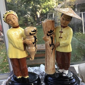 Peut inclure: Deux figurines en céramique vintage représentant des personnes asiatiques en vêtements traditionnels. La figurine de gauche tient un vase brun et noir. La figurine de droite tient une tige de bambou brun et blanc. Les deux figurines sont posées sur des socles noirs.