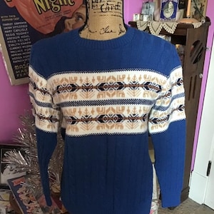 May include: A blue knitted sweater with a white, brown, and black geometric pattern. The sweater has a crew neck and long sleeves.
